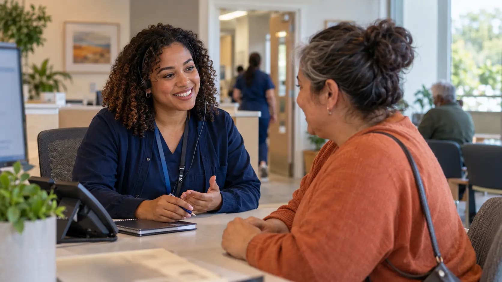 Reception colleague holding a pen while talking with a visitor at a clinic desk
