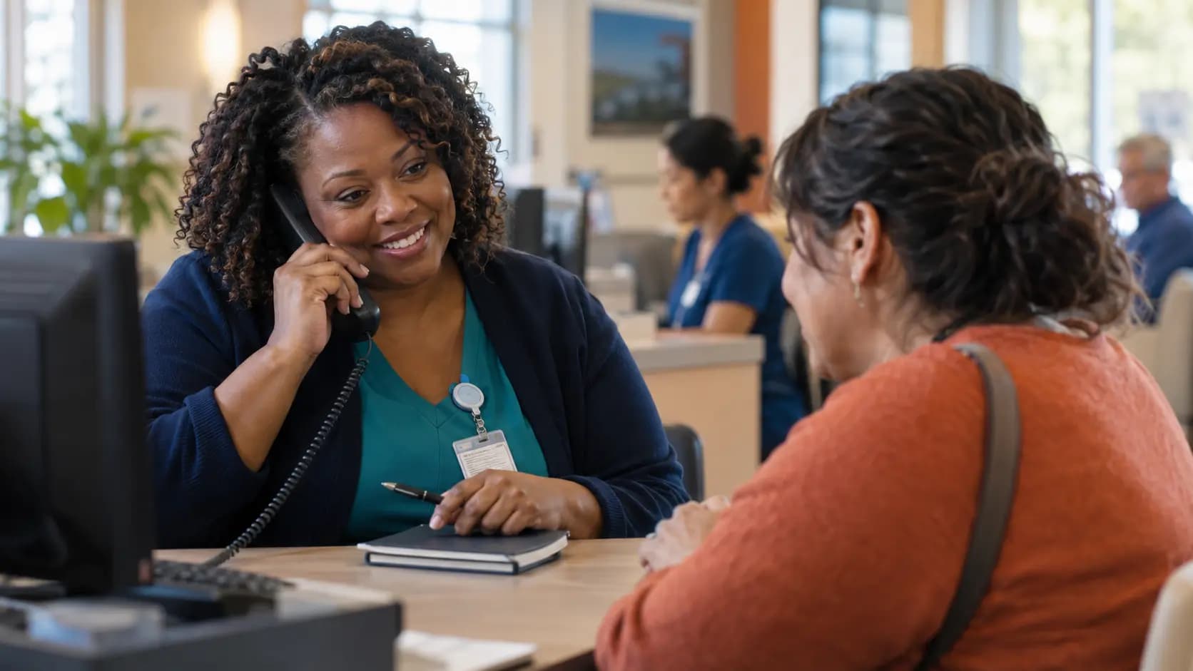Reception colleague using a telephone handset while assisting a visitor at a clinic desk