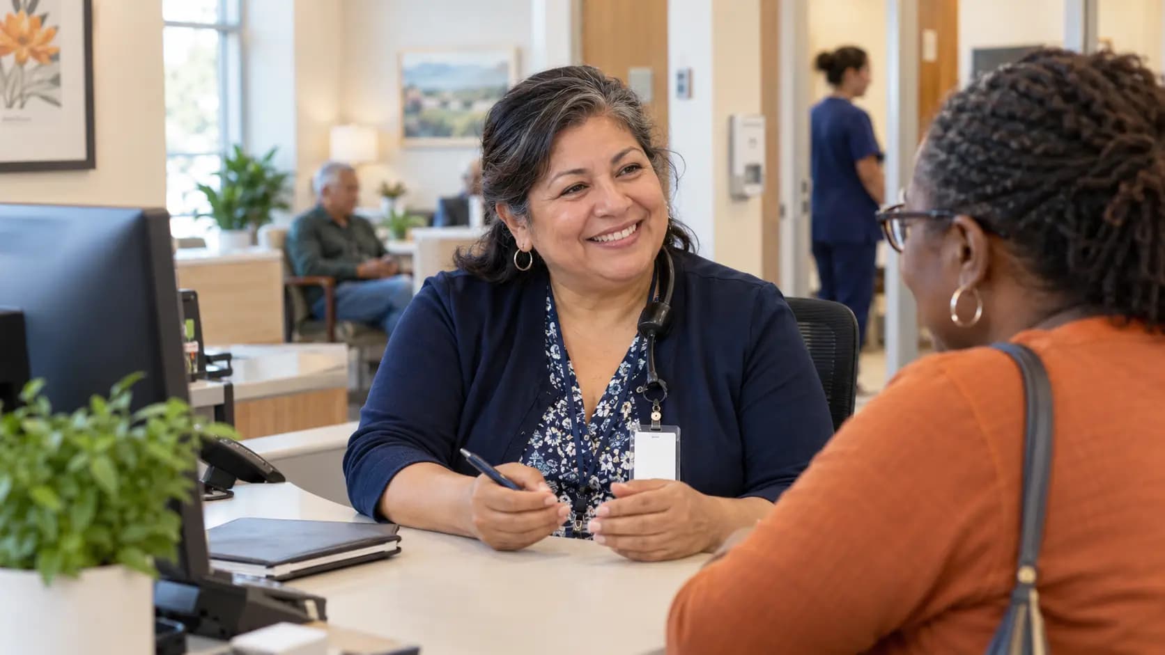 Reception colleague holding a pen and talking with a visitor across a clinic desk