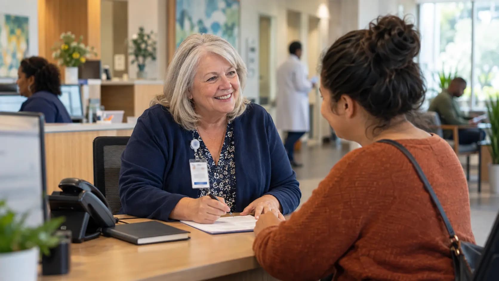 Reception colleague writing on a form while speaking with a visitor at a clinic desk